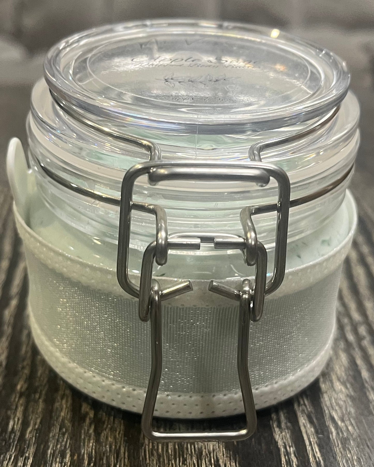 Clear round jar with a sticker on top. You see through to the jar to green body butter. You see the side of the jar, which is covered in a sparkly white ribbon.