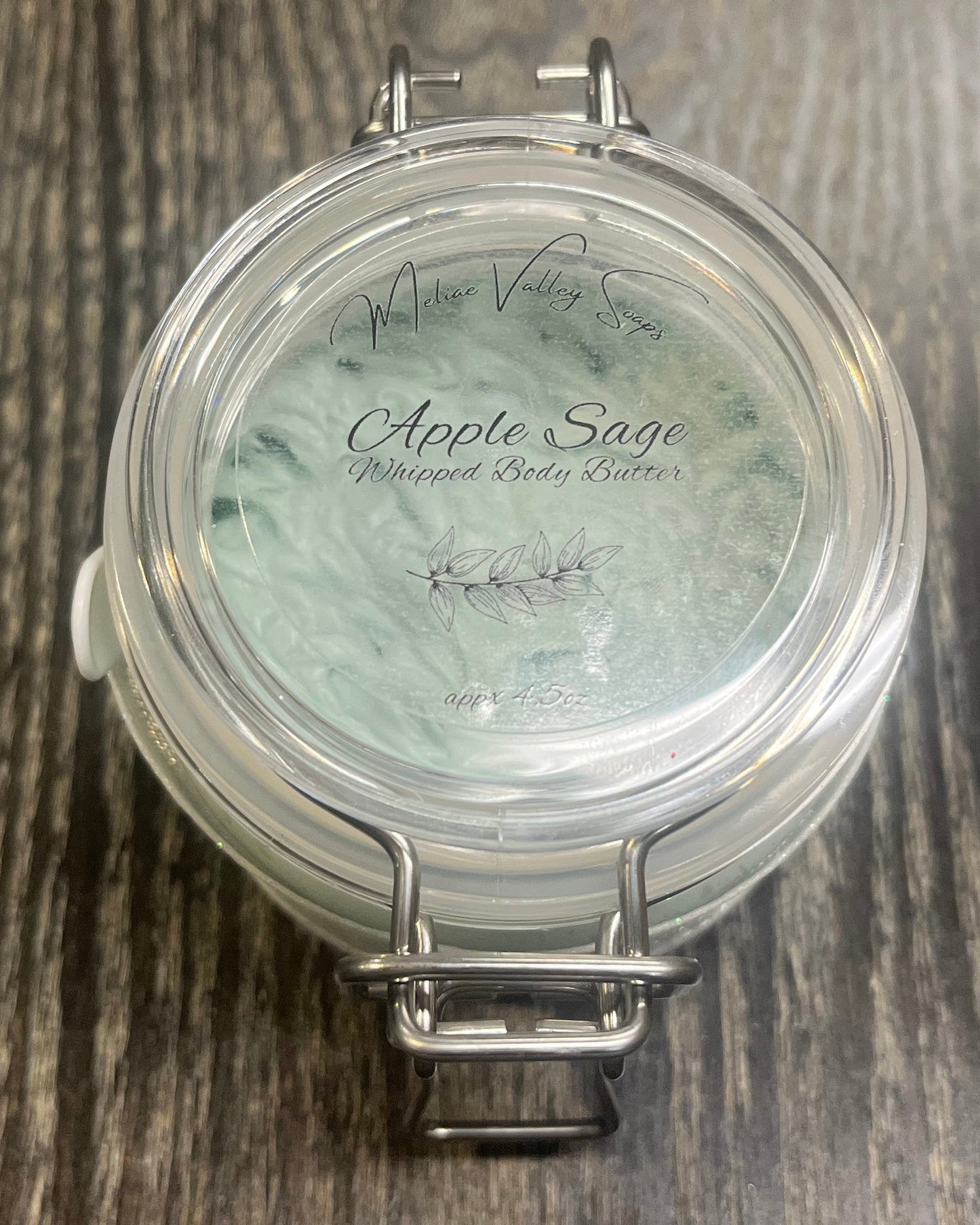 Clear round jar with a sticker on top. You see through to the jar of green body butter. 