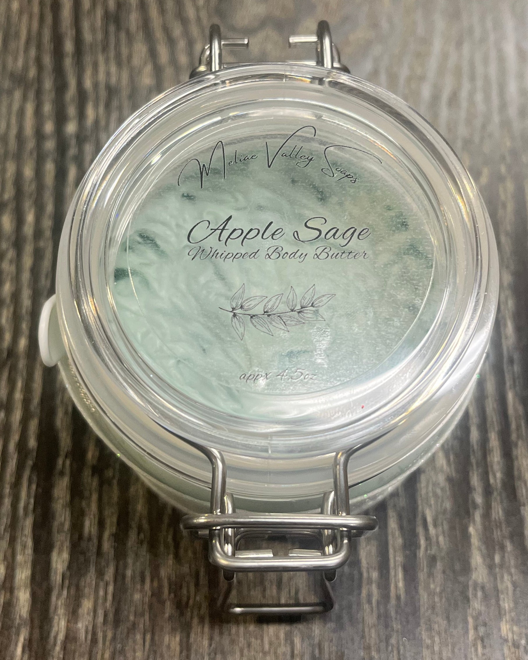 Clear round jar with a sticker on top. You see through to the jar of green body butter. 