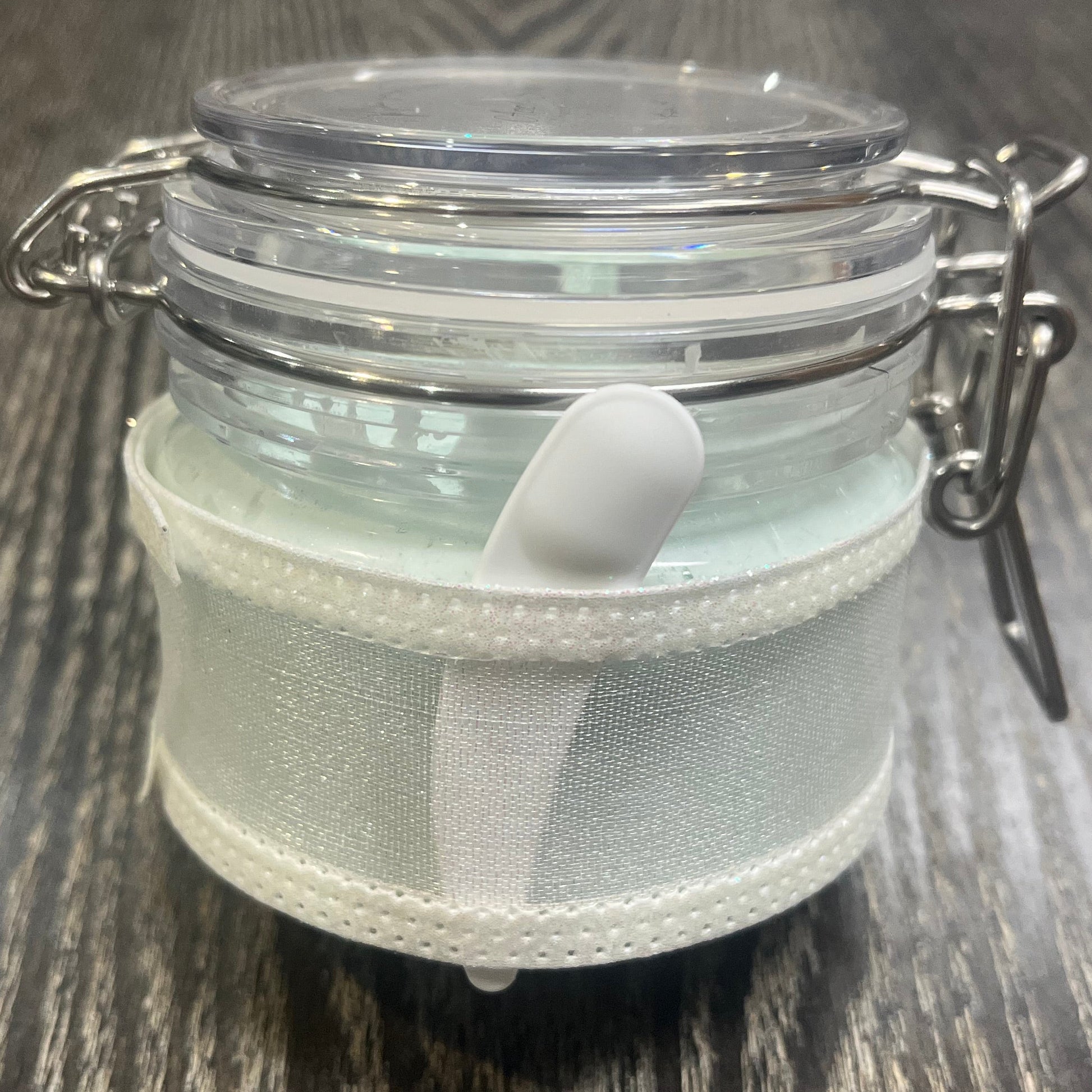 Clear round jar with a sticker on top. You see through to the jar to green body butter. You see the side of the jar, which is covered in a sparkly white ribbon. Between the jar and the ribbon a mini-makeup spatula. 