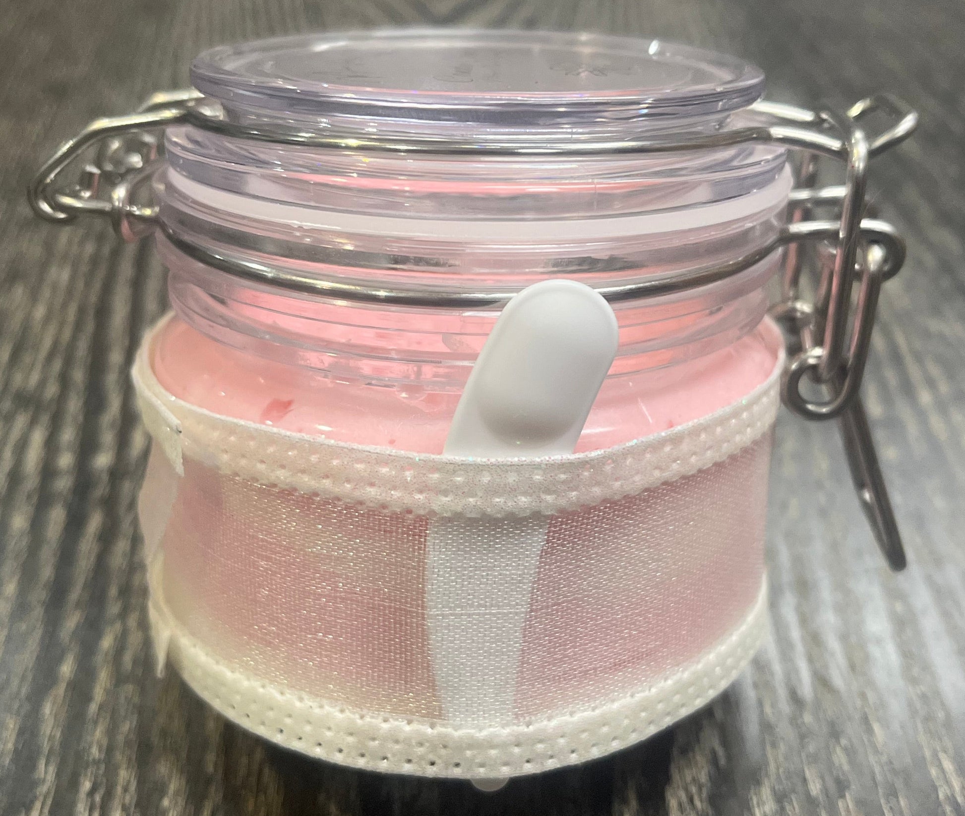 Clear round jar with a sticker on top. You see through to the jar to pink body butter. You see the side of the jar, which is covered in a sparkly white ribbon. Between the jar and the ribbon a mini-makeup spatula. 