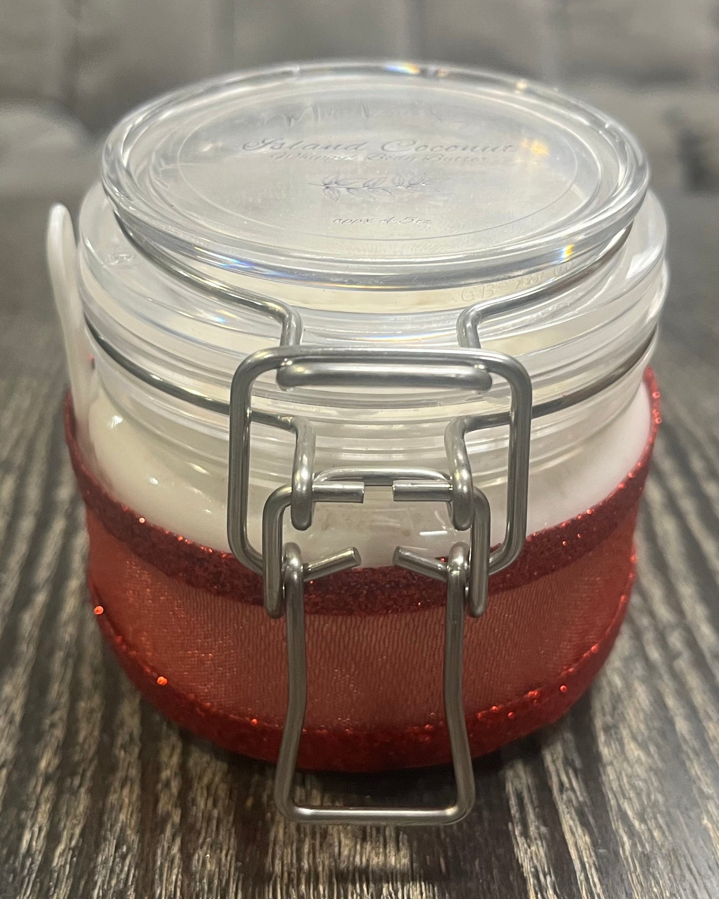 Clear round jar with a sticker on top. You see through to the jar to white body butter. You see the side of the jar, which is covered in a sparkly red ribbon.
