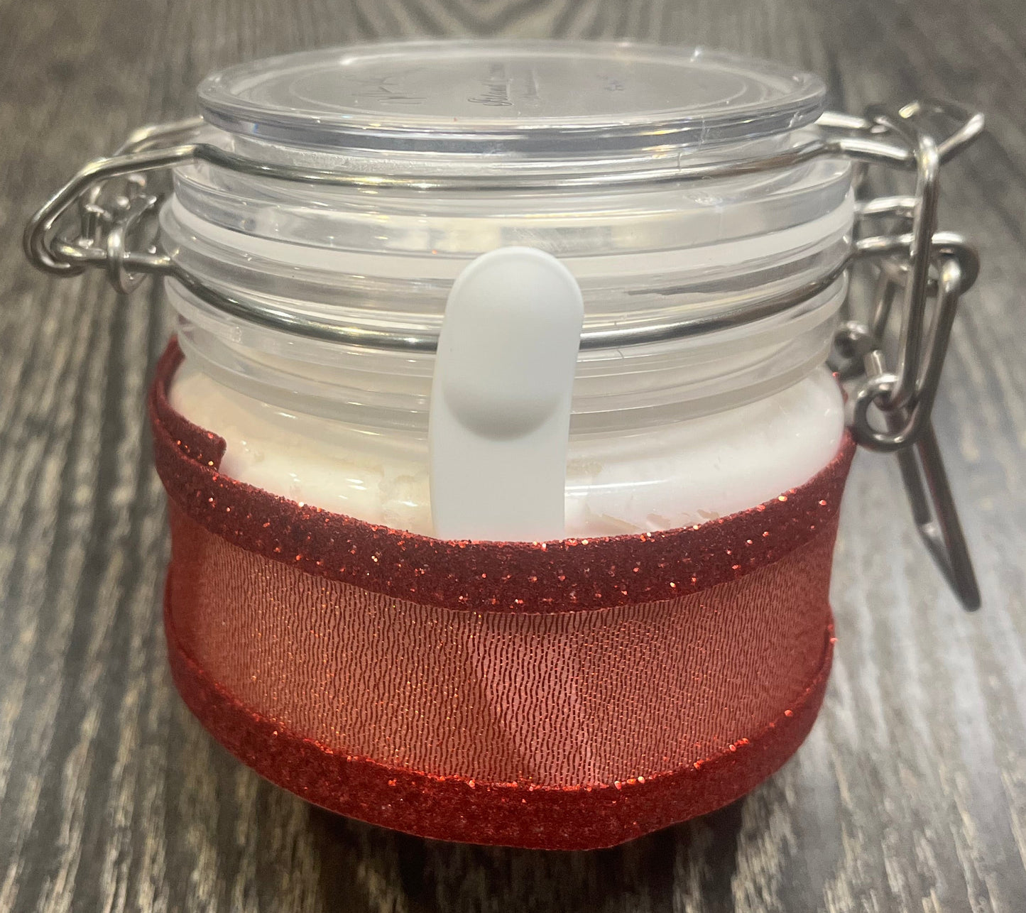 Clear round jar with a sticker on top. You see through to the jar to white body butter. You see the side of the jar, which is covered in a sparkly red ribbon. Between the jar and the ribbon a mini-makeup spatula. 