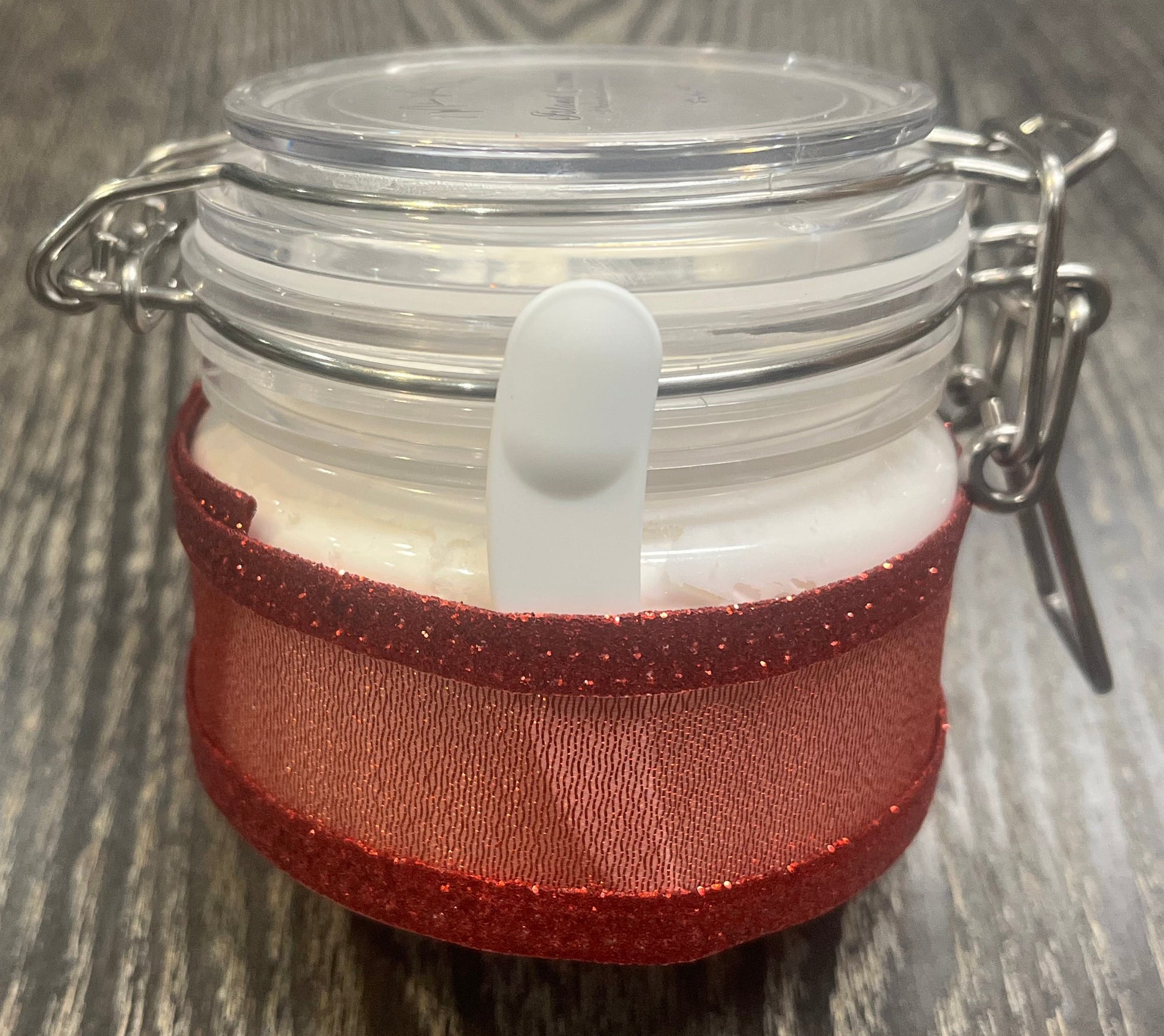 Clear round jar with a sticker on top. You see through to the jar to white body butter. You see the side of the jar, which is covered in a sparkly red ribbon. Between the jar and the ribbon a mini-makeup spatula. 