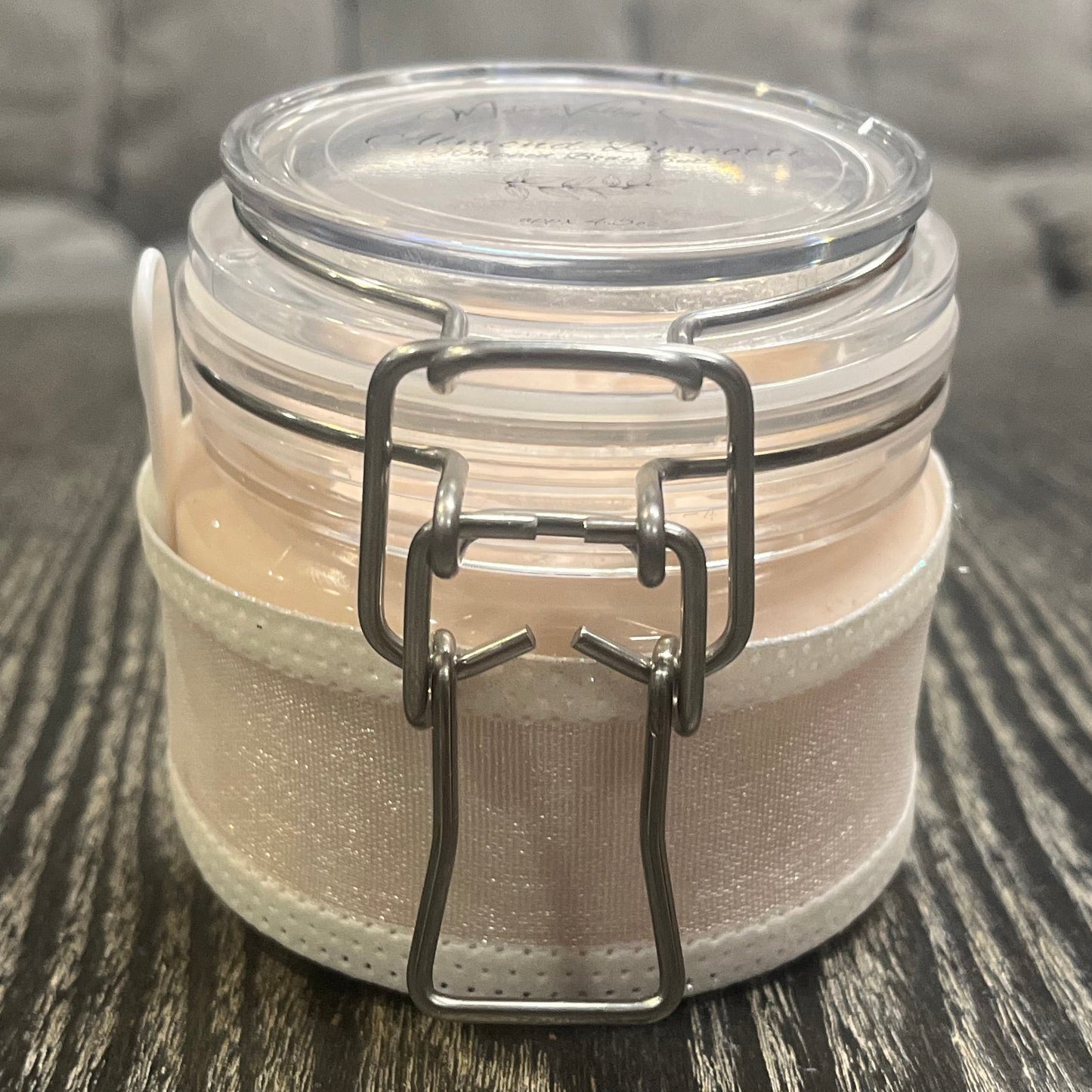 Clear round jar with a sticker on top. You see through to the jar to bronze body butter. You see the side of the jar, which is covered in a sparkly white ribbon.