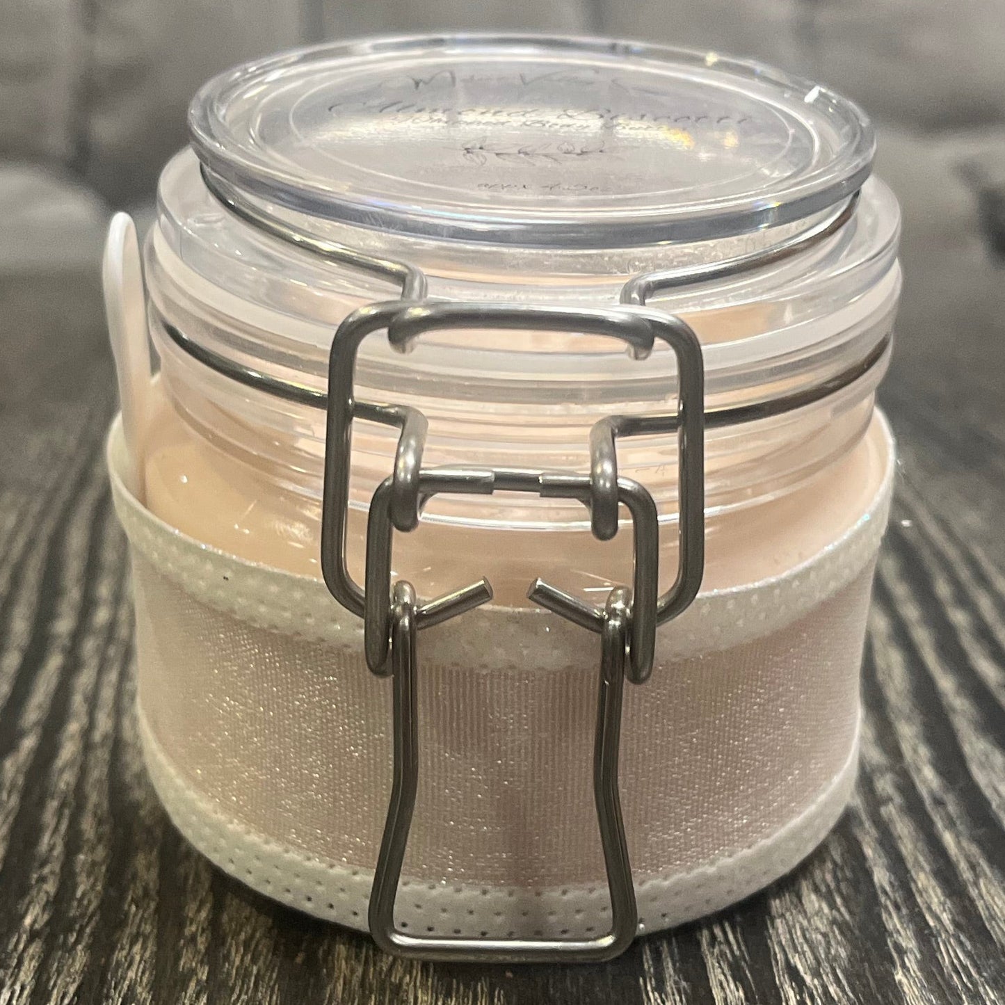 Clear round jar with a sticker on top. You see through to the jar to bronze body butter. You see the side of the jar, which is covered in a sparkly white ribbon.