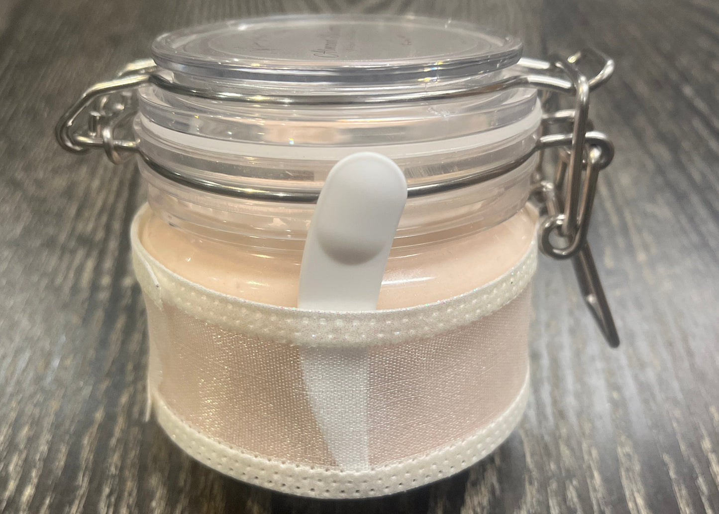 Clear round jar with a sticker on top. You see through to the jar to bronze body butter. You see the side of the jar, which is covered in a sparkly white ribbon. Between the jar and the ribbon a mini-makeup spatula. 