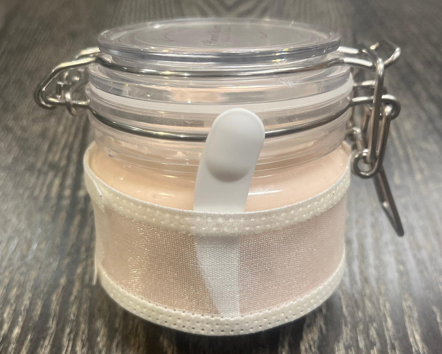 Clear round jar with a sticker on top. You see through to the jar to bronze body butter. You see the side of the jar, which is covered in a sparkly white ribbon. Between the jar and the ribbon a mini-makeup spatula. 