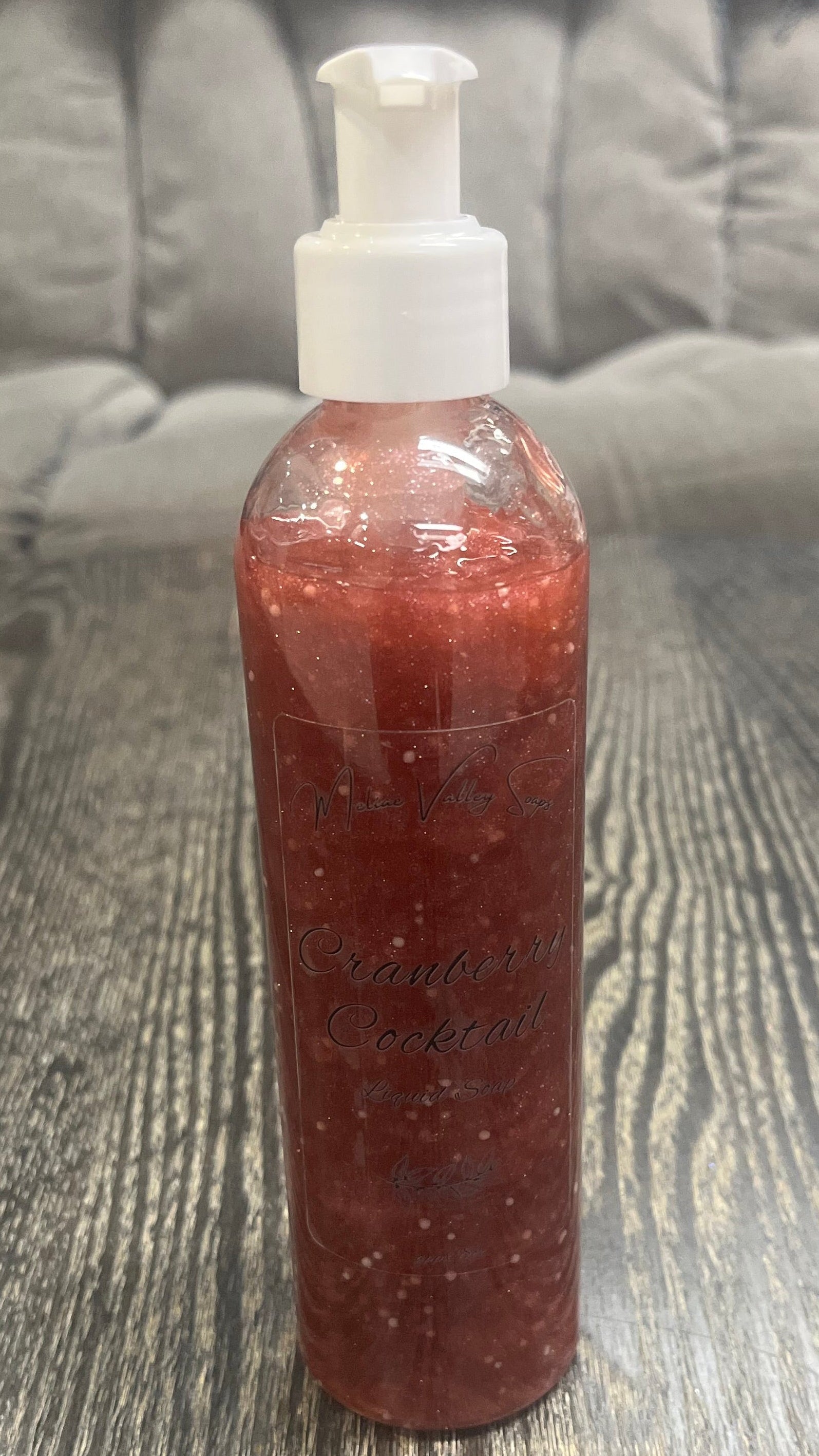 A clear pump bottle with a white nozzle full of red liquid with white beads on a textured surface. 