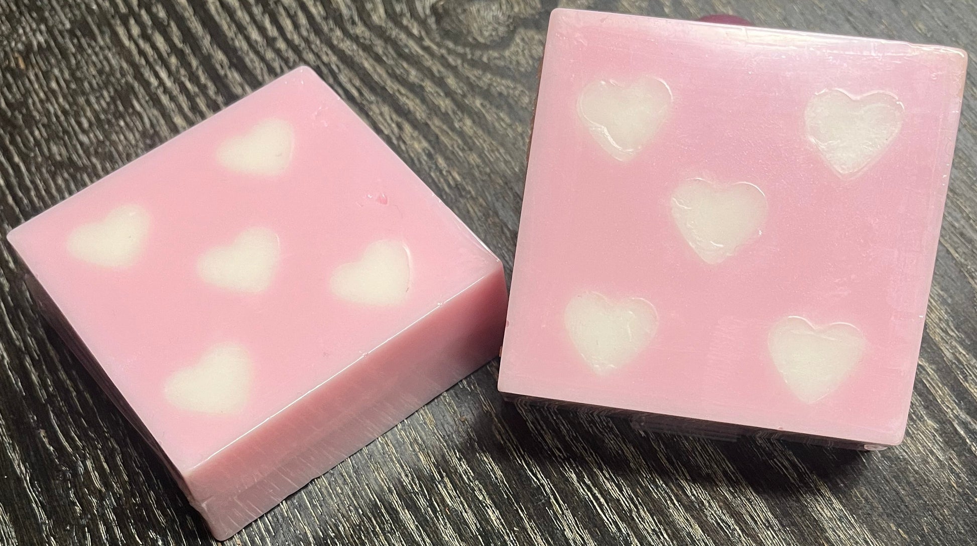 Two pink soap bars with white heart patterns on a dark wooden surface.