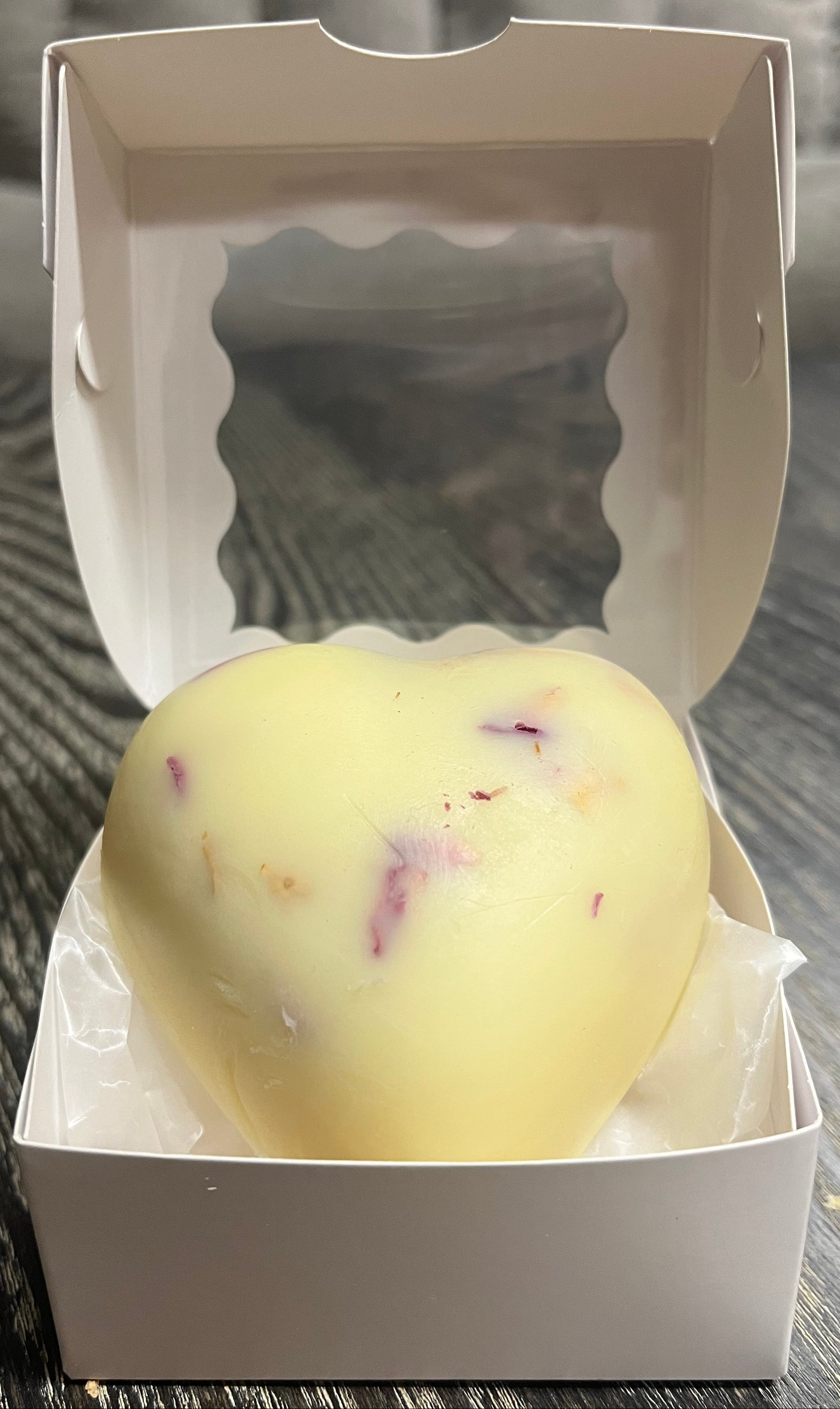 Cream colored, heart-shaped, lotion bar in an open white box on a wooden surface.