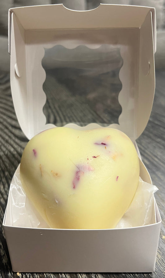 Cream colored, heart-shaped, lotion bar in an open white box on a wooden surface.