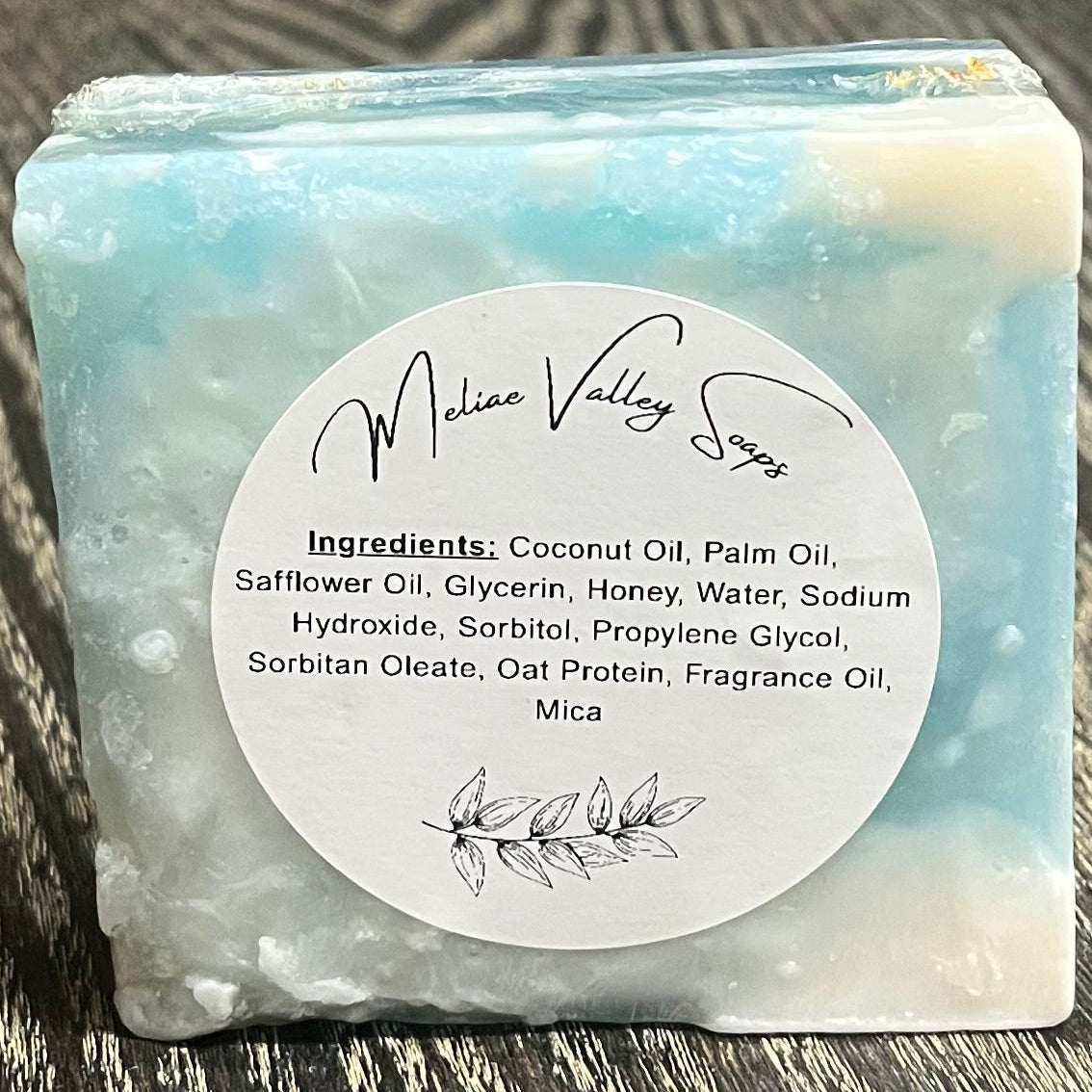 A multi colored, clear, white, and blue soap bar with a white ingredients label. It is upright on its side, showing the bottom of the bar. 