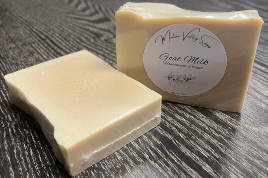 Two bars of beige soap, one is upright with a label while the other is face down, showing the back of the bar on a wooden surface. 