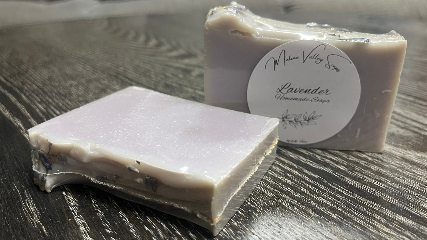 Two bars of lavender soap, one upright with a white label, the other shows the textured top of the bars with lavender flowers. 
