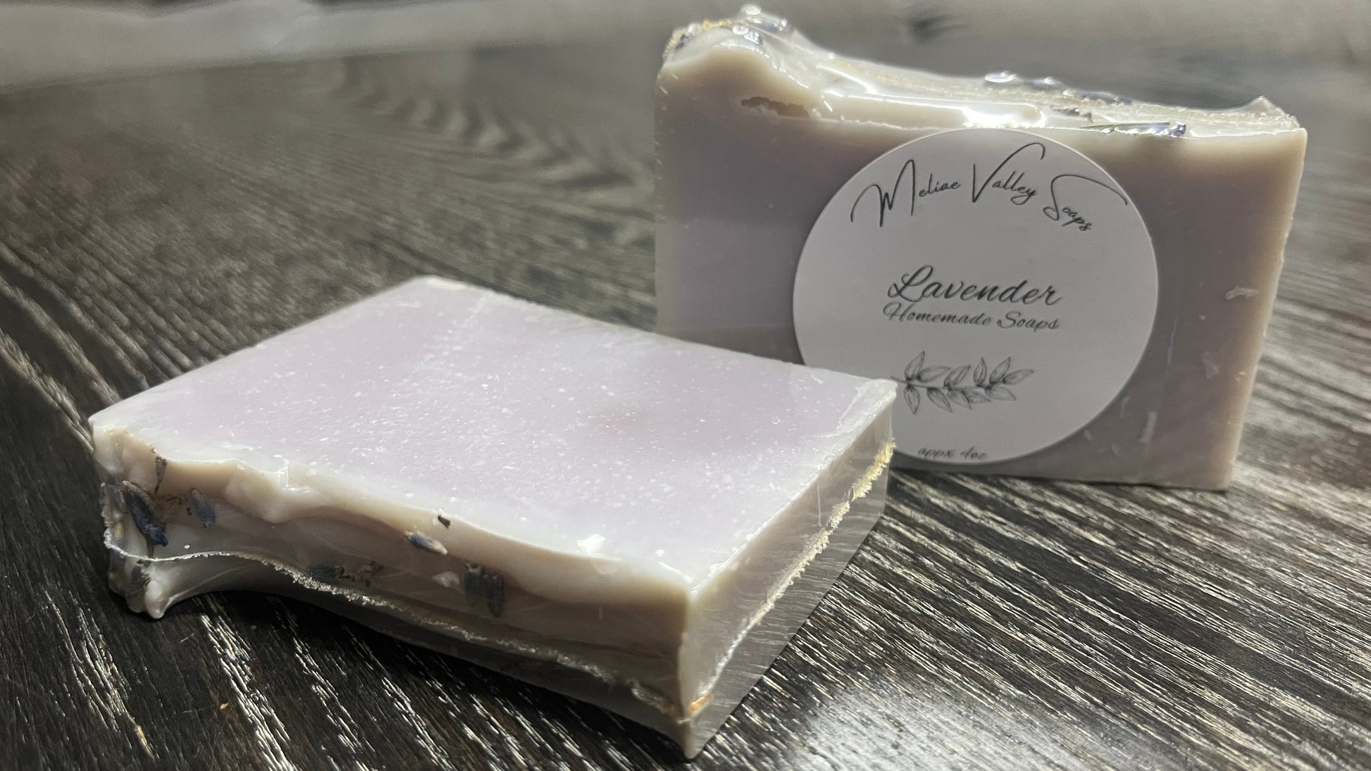 Two bars of lavender soap, one upright with a white label, the other shows the textured top of the bars with lavender flowers. 