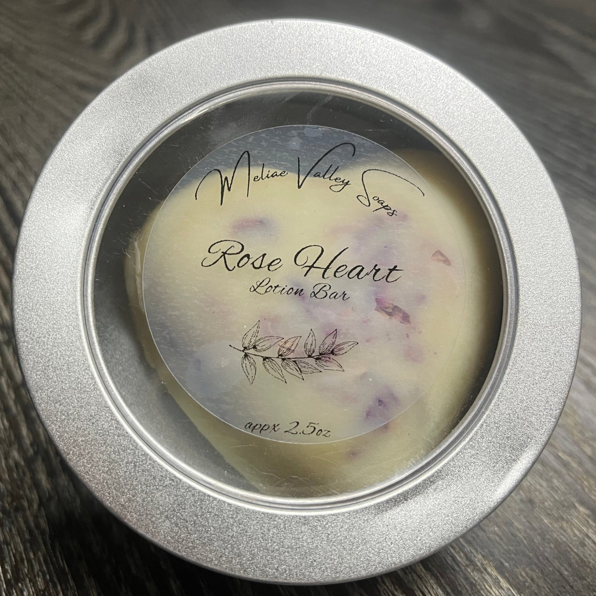 Heart-shaped white lotion bar with visible rose petals in a tin with a clear label.
