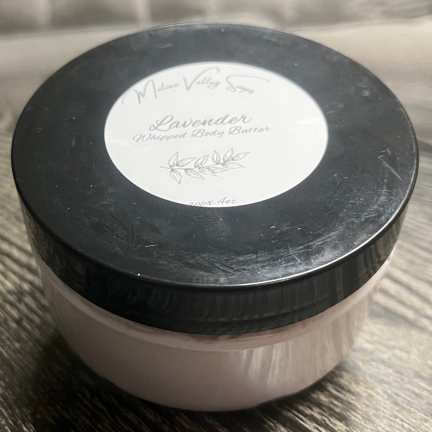 Jar of lavender body butter with a black lid and a white label on a wooden surface.