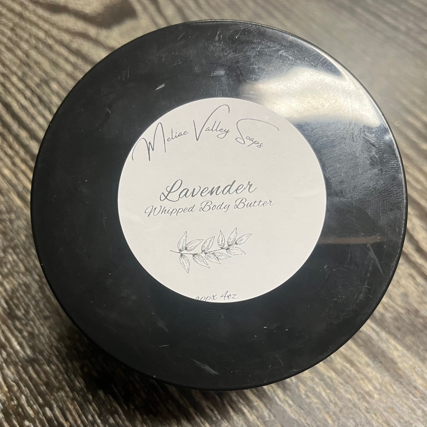 A jar of lavender whipped body butter with a black lid and a white label on a wooden surface. 