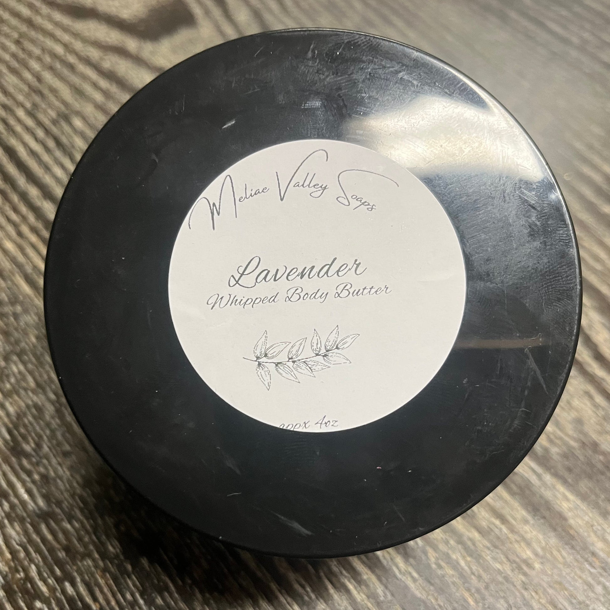 A jar of lavender whipped body butter with a black lid and a white label on a wooden surface. 