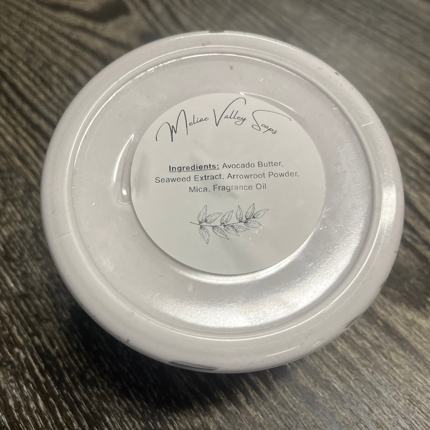 A clear jar of whipped body butter with a white ingredients label on a wooden surface. 