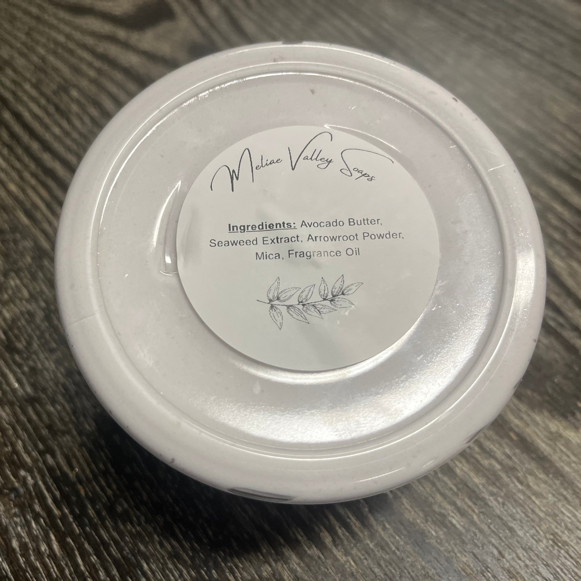 A clear jar of whipped body butter with a white ingredients label on a wooden surface. 
