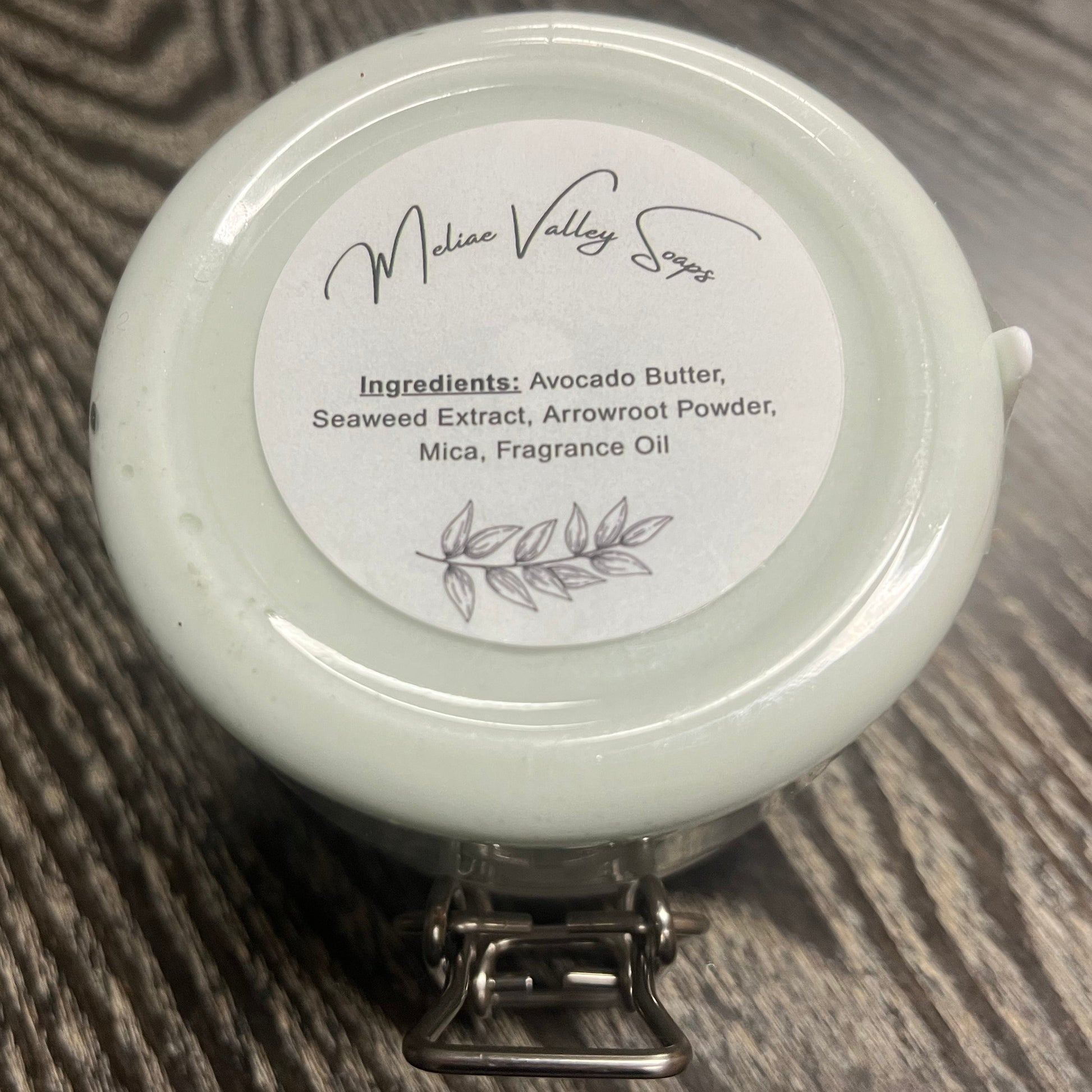 Jar of green body butter with a white ingredients label on a wooden surface. 