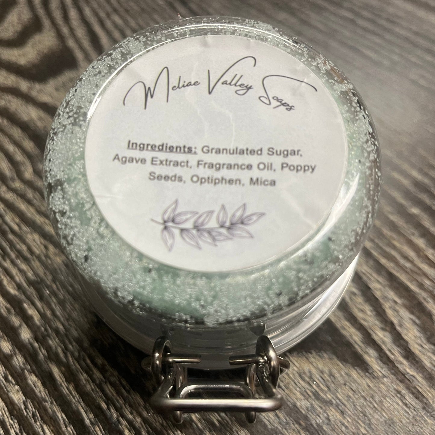 Jar of green sugar scrub with a white ingredients label on a wooden surface. 