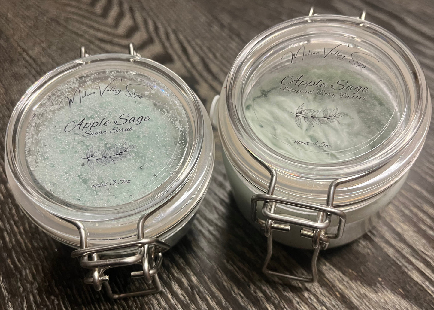 Two plastic jars with lids on a wooden surface. The smaller jar on the left holds green sugar body scrub. The bigger right jar holds green whipped body butter. 