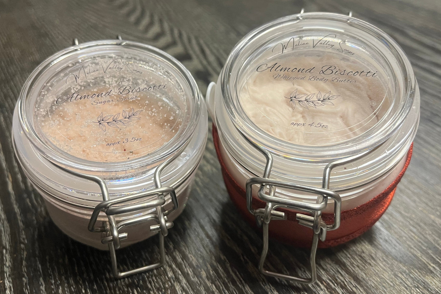 Two plastic jars with lids on a wooden surface. The smaller jar on the left holds tan sugar body scrub. The bigger right jar holds tan whipped body butter. 