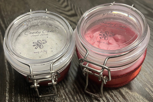 Two plastic jars with lids on a wooden surface. The smaller jar on the left holds white sugar body scrub. The bigger right jar holds pink whipped body butter. 