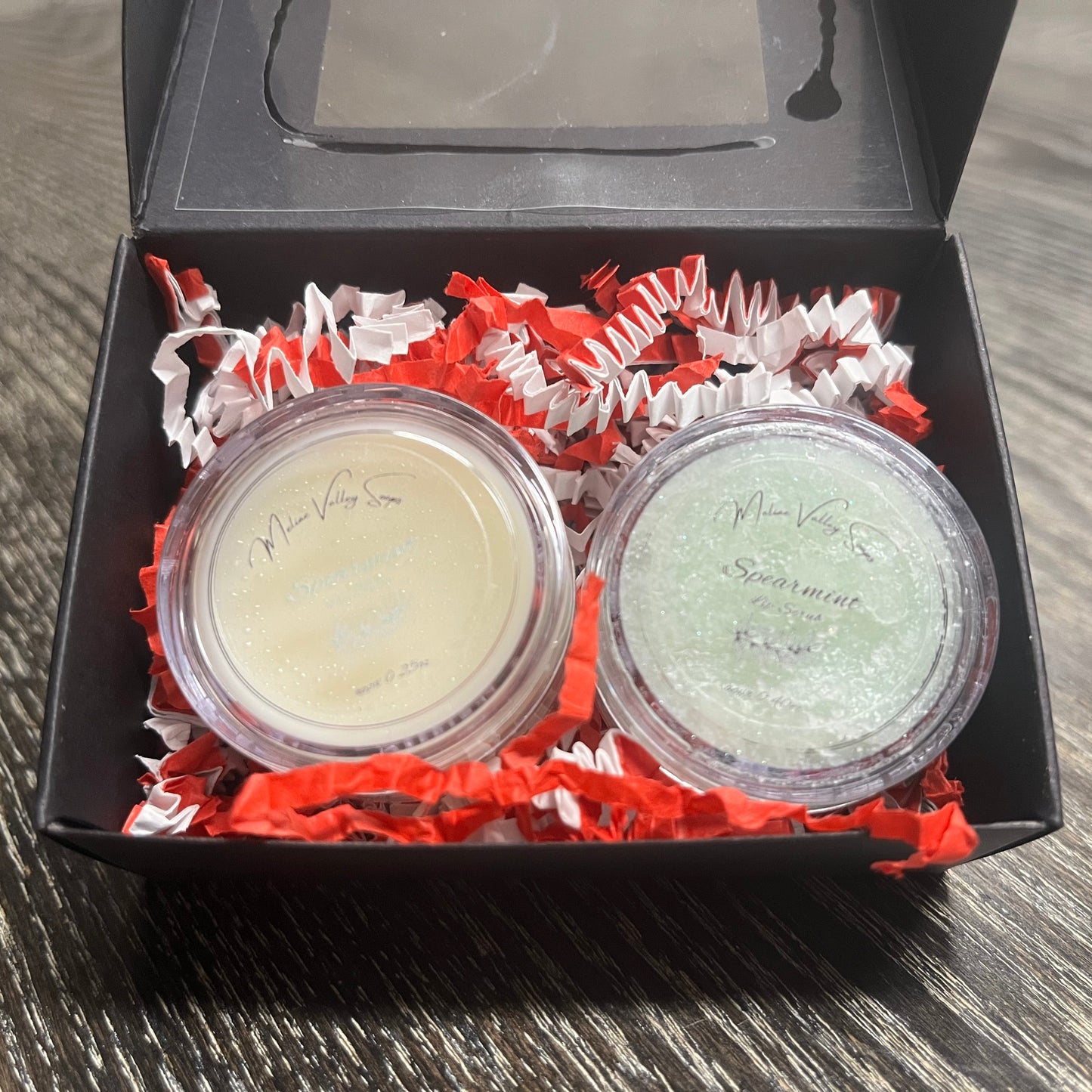 Two lip pots in a black box with red crinkled paper on a wooden surface. The lip pot on the left is lip balm, and the lip pot on the right is green colored lip scrub. 