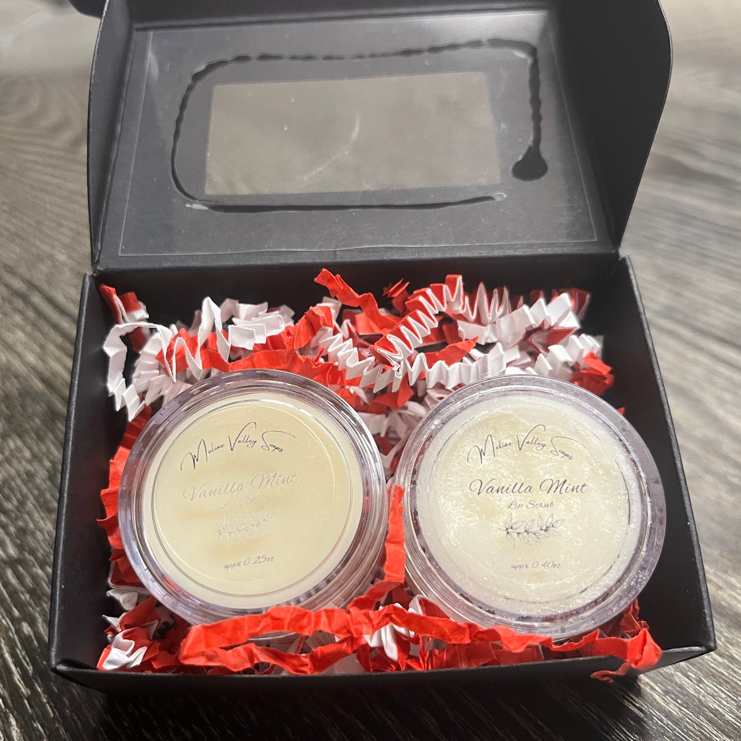 Two lip pots in a black box with red crinkled paper on a wooden surface. The lip pot on the left is lip balm, and the lip pot on the right is white colored lip scrub. 