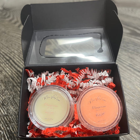 Two lip pots in a black box with red crinkled paper on a wooden surface. The lip pot on the left is lip balm, and the lip pot on the right is orange colored lip scrub. 