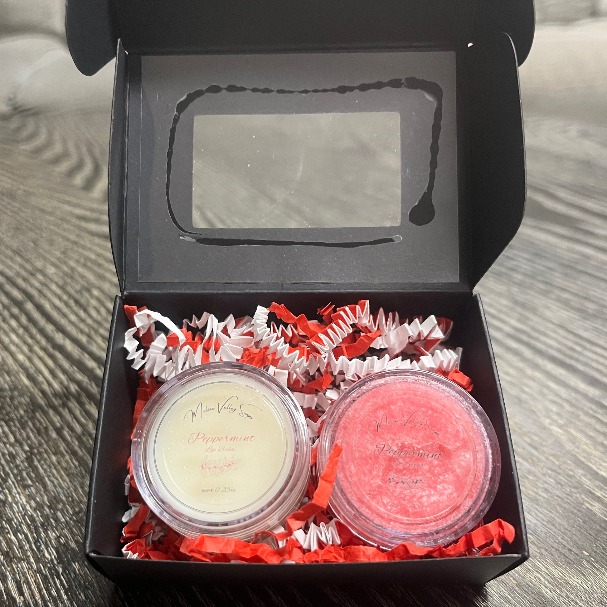 Two lip pots in a black box with red crinkled paper on a wooden surface. The lip pot on the left is lip balm, and the lip pot on the right is red colored lip scrub. 
