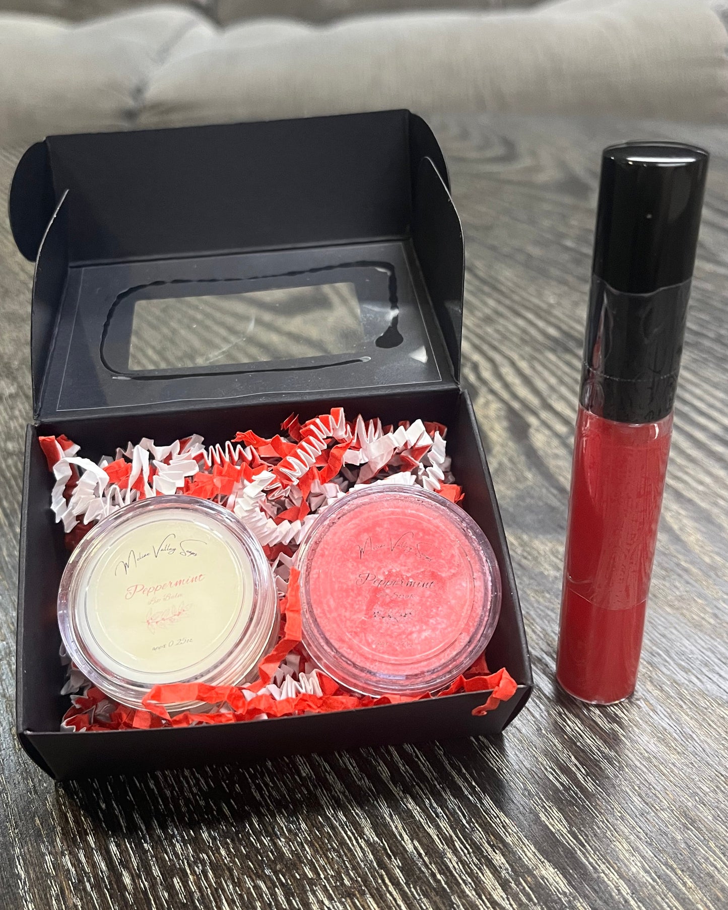 Two lip pots in a black box with red crinkled paper on a wooden surface. The lip pot on the left is lip balm, and the lip pot on the right is red colored lip scrub. On the right of the box there is a red tube of lip gloss. 