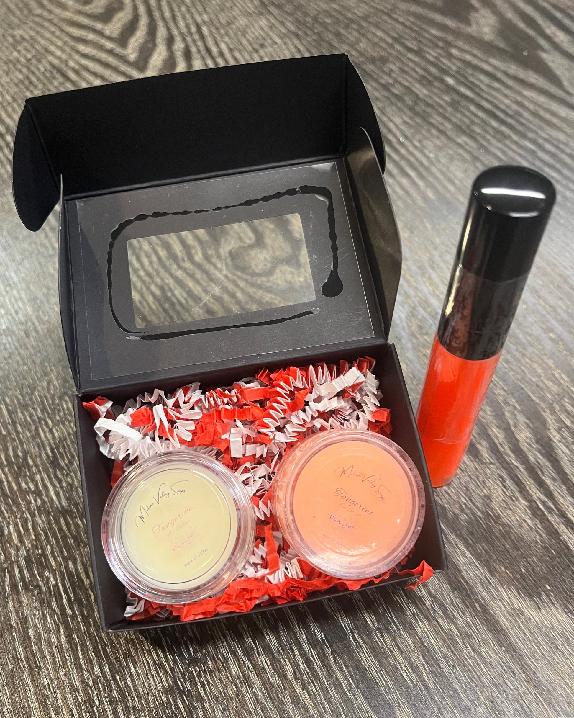 Two lip pots in a black box with red crinkled paper on a wooden surface. The lip pot on the left is lip balm, and the lip pot on the right is orange colored lip scrub. On the right of the box there is a bright orange tube of lip gloss. 