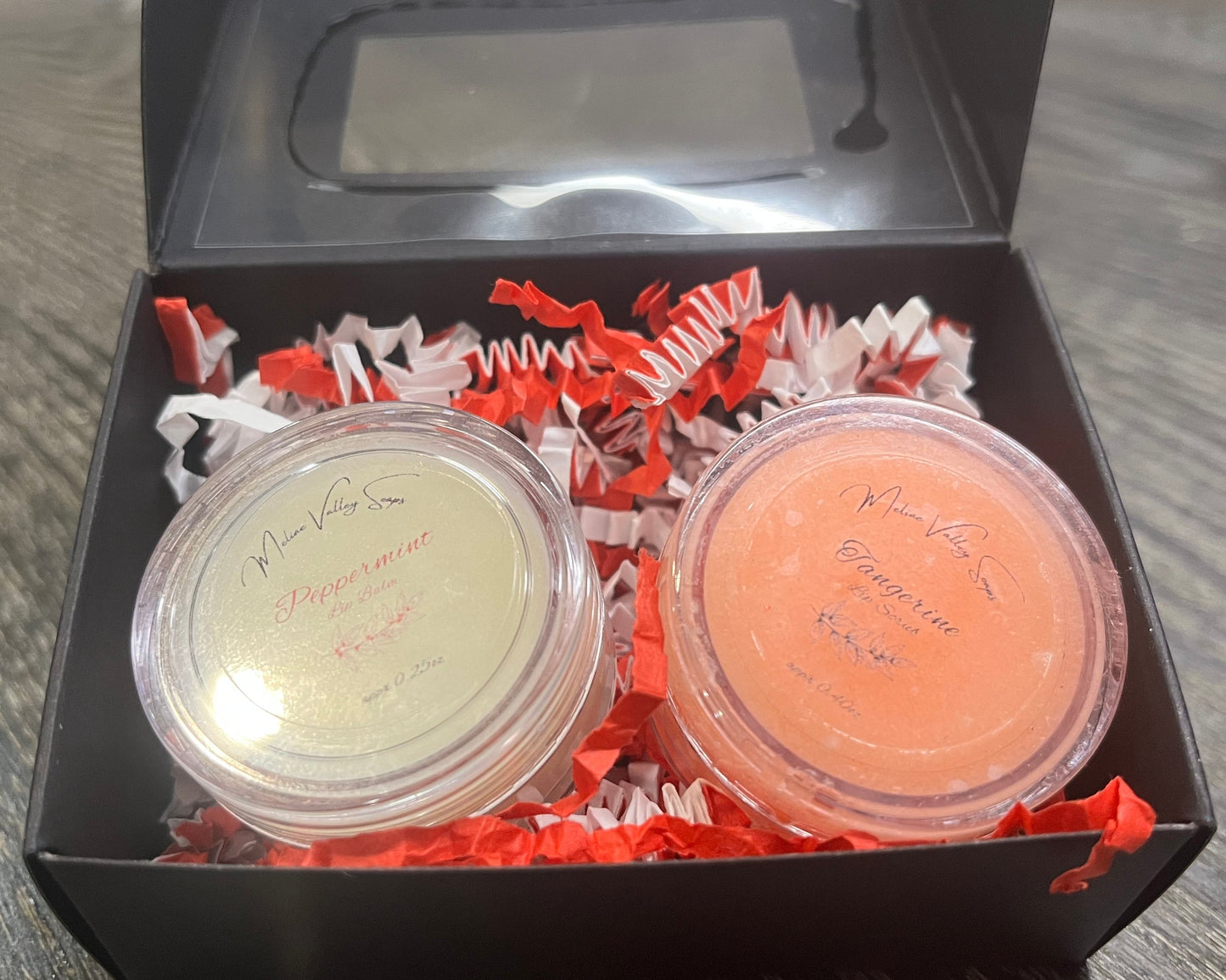 Two lip pots in a black box with red crinkled paper on a wooden surface. The lip pot on the left is lip balm, and the lip pot on the right is orange colored lip scrub. 