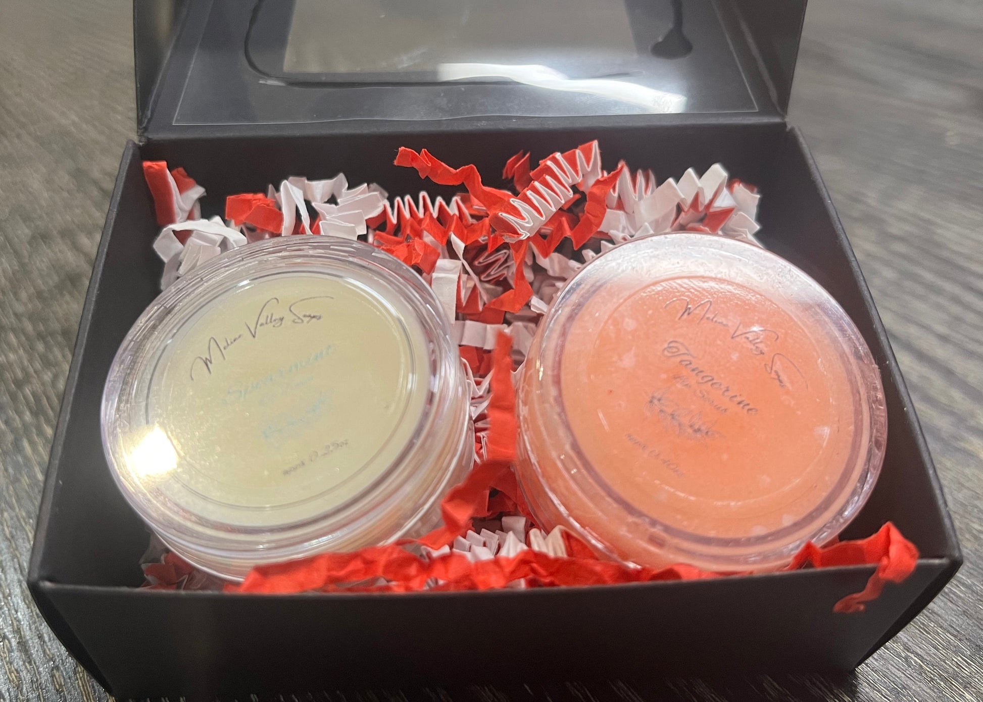 Two lip pots in a black box with red crinkled paper on a wooden surface. The lip pot on the left is lip balm, and the lip pot on the right is orange colored lip scrub. 