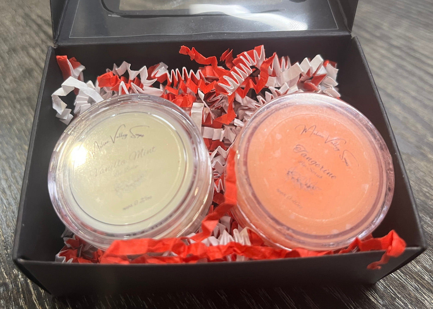 Two lip pots in a black box with red crinkled paper on a wooden surface. The lip pot on the left is lip balm, and the lip pot on the right is orange colored lip scrub. 