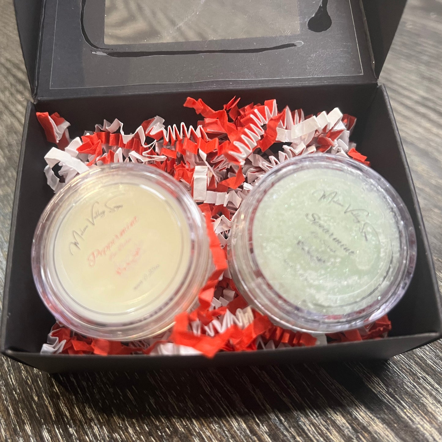 Two lip pots in a black box with red crinkled paper on a wooden surface. The lip pot on the left is lip balm, and the lip pot on the right is green colored lip scrub. 