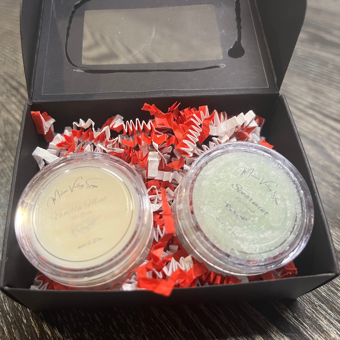 Two lip pots in a black box with red crinkled paper on a wooden surface. The lip pot on the left is lip balm, and the lip pot on the right is green colored lip scrub. 