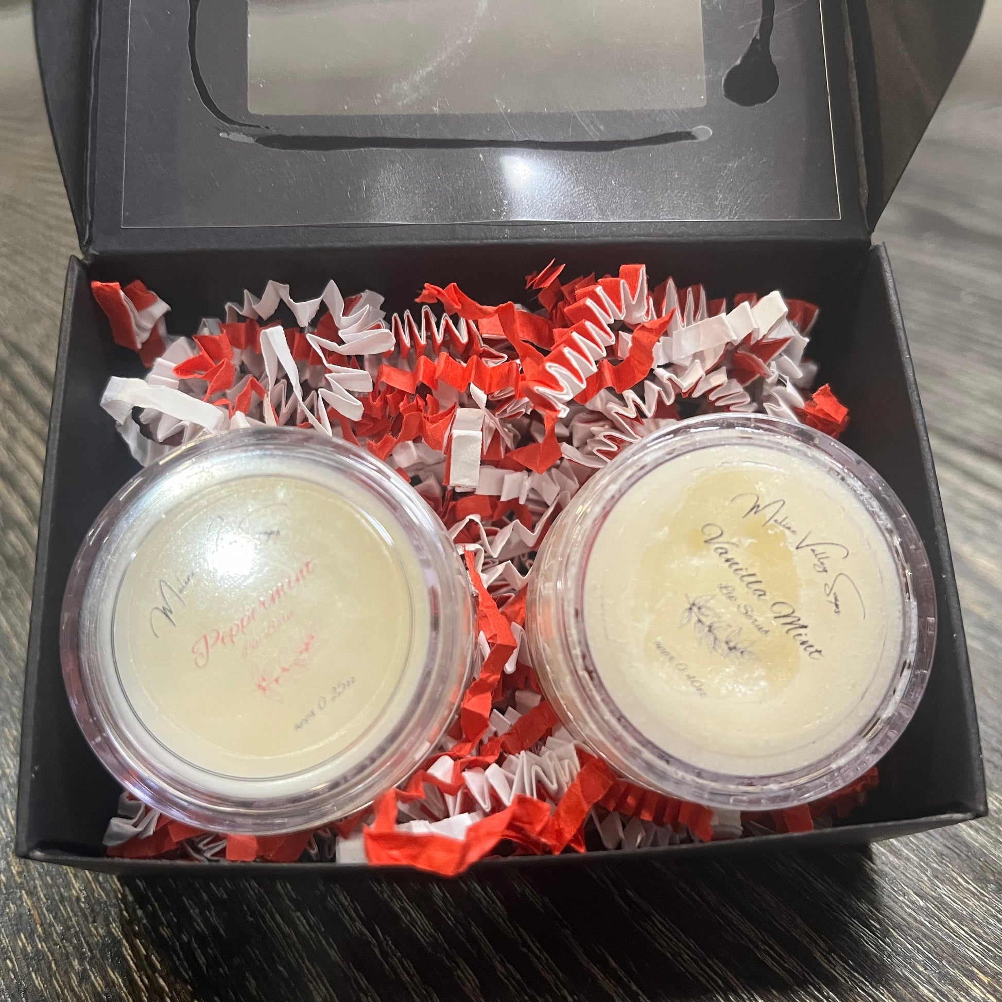 Two lip pots in a black box with red crinkled paper on a wooden surface. The lip pot on the left is lip balm, and the lip pot on the right is white colored lip scrub. 