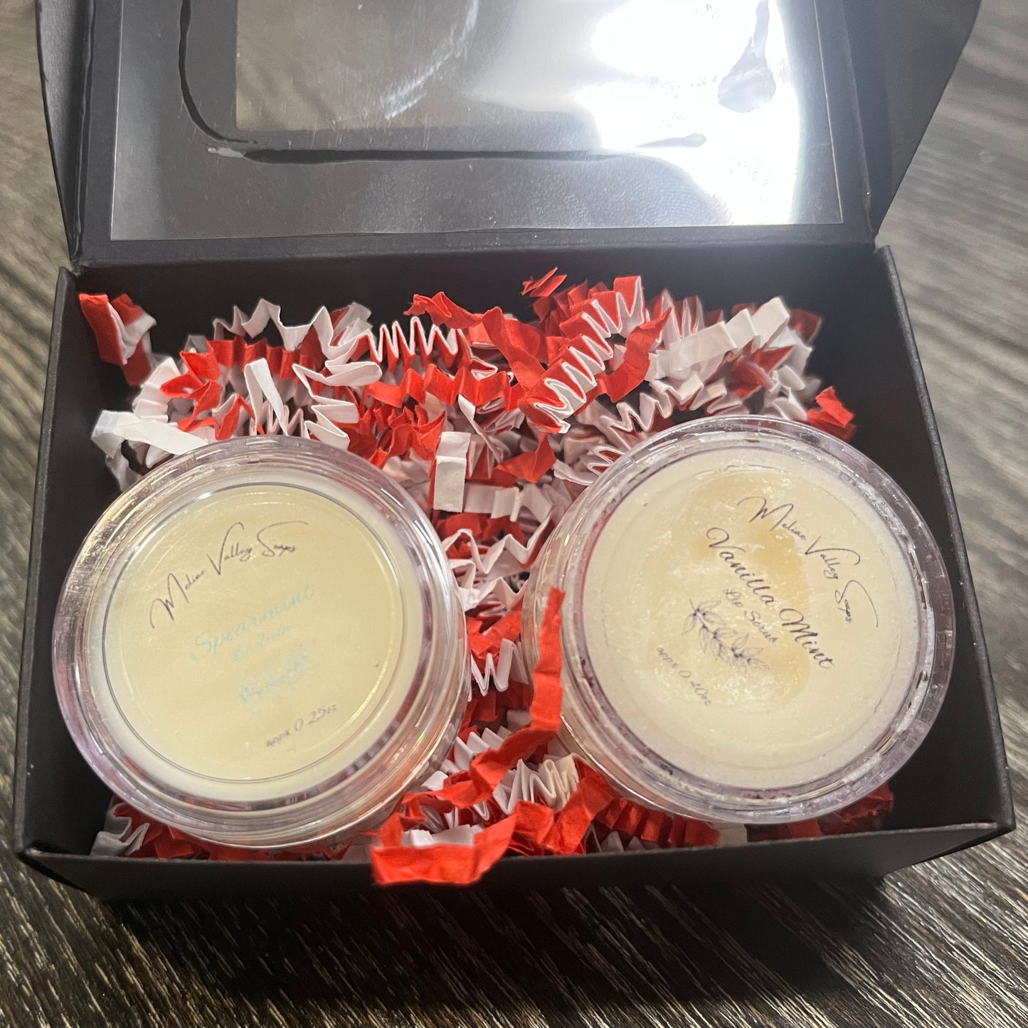 Two lip pots in a black box with red crinkled paper on a wooden surface. The lip pot on the left is lip balm, and the lip pot on the right is white colored lip scrub. 
