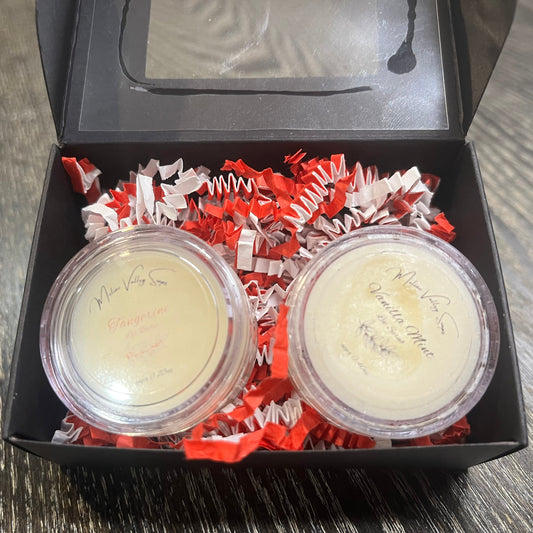 Two lip pots in a black box with red crinkled paper on a wooden surface. The lip pot on the left is lip balm, and the lip pot on the right is white colored lip scrub. 