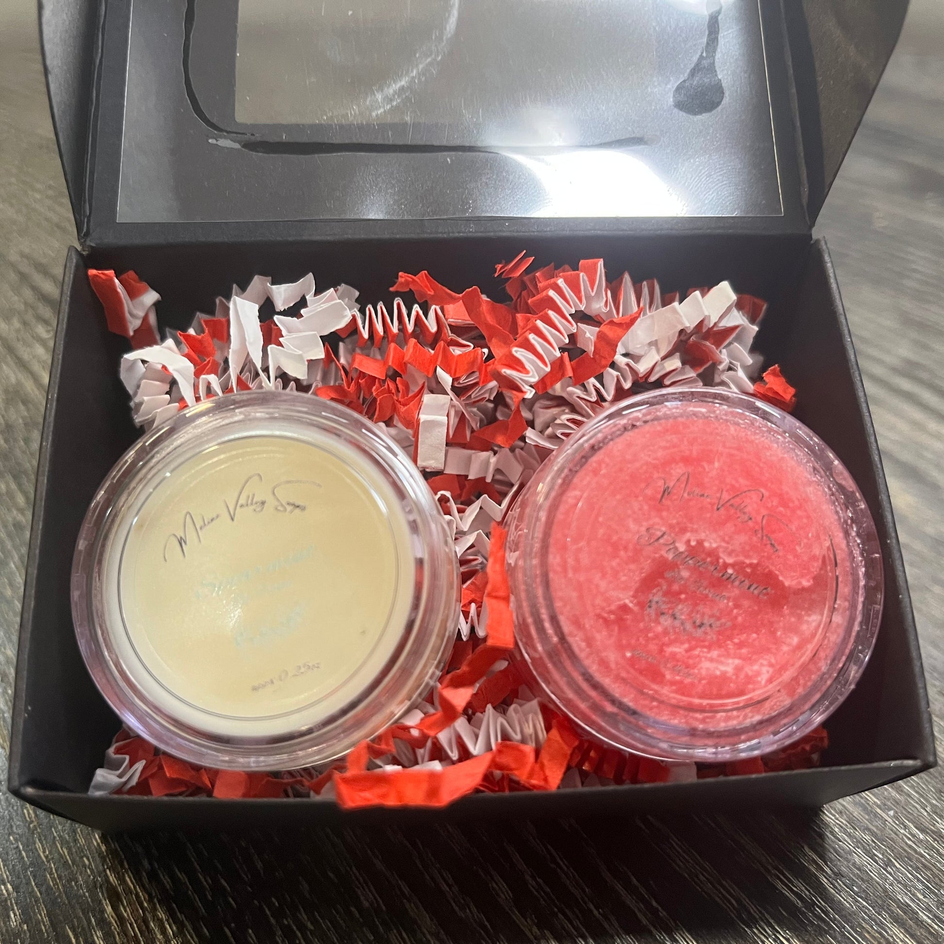 Two lip pots in a black box with red crinkled paper on a wooden surface. The lip pot on the left is lip balm, and the lip pot on the right is red colored lip scrub. 
