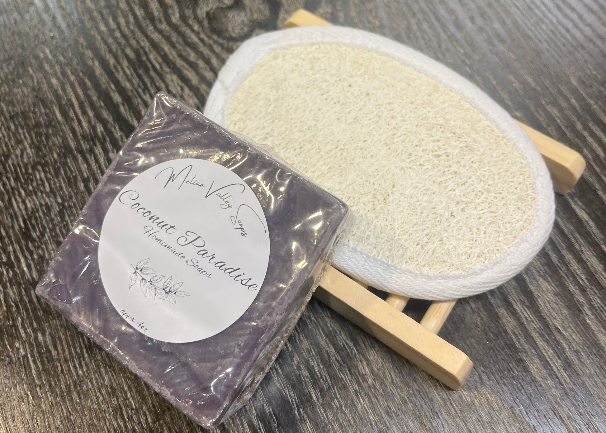 A purple bar of soap with a wooden stand and a loofah pad on a dark wooden surface. 