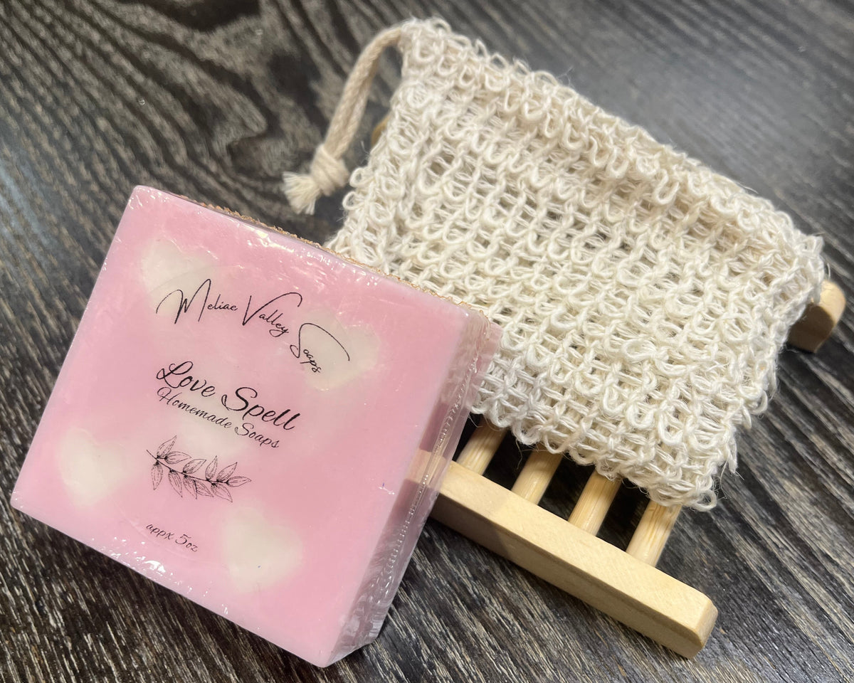 A pink soap bar with white hearts and a white sticker leans against an exfoliating bag and a soap bar holder. 