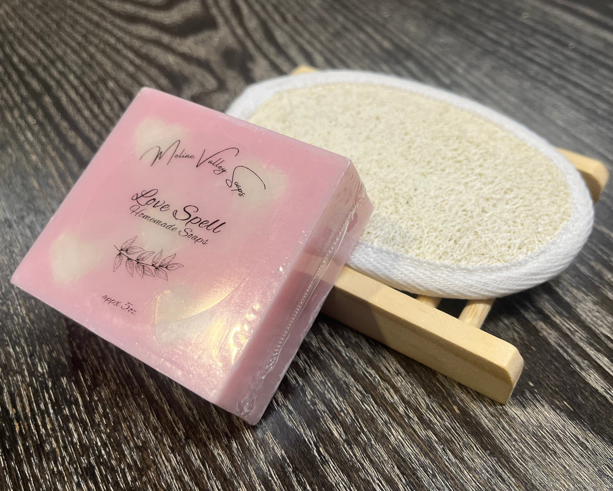 A pink soap bar with hearts and a white sticker leans against a loofah pad and a soap bar holder.