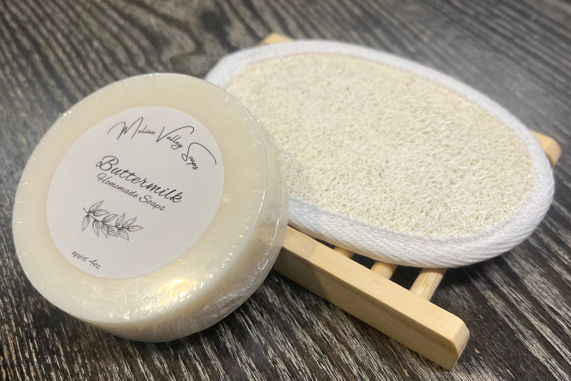 A round white soap with a wooden stand and a loofah pad on a dark wooden surface. 